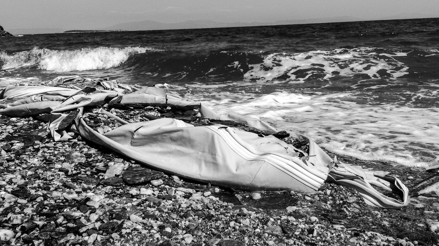 A deflated inflatable boat rests on the rocky shore, echoing the tumultuous power of nature as waves crash in the background—a reminder of our fragile relationship with climate and environment. | Jacob Warn