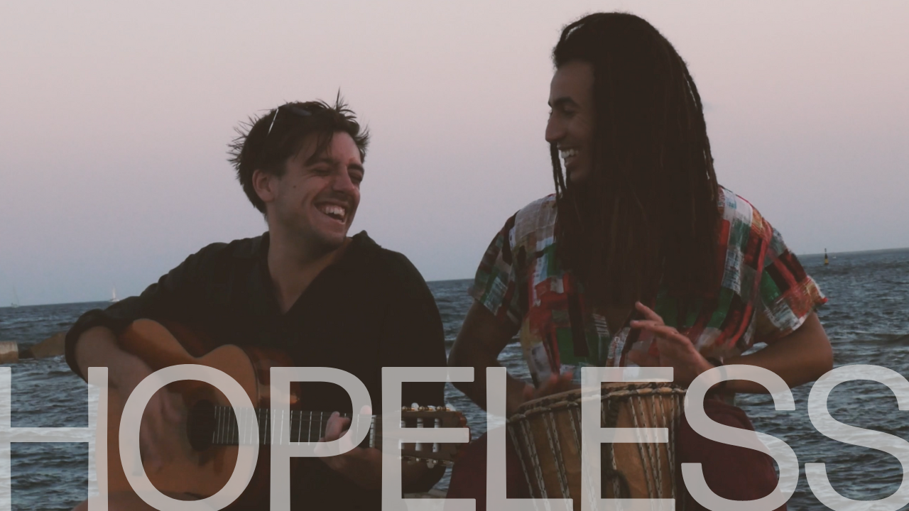 Hopeless Cover Art (1280 × 720px) (1) Taha and Jacob smile while playing instruments by the sea at dusk, with "Hopeless," their new song's title, prominently displayed in large text at the bottom. | Jacob Warn
