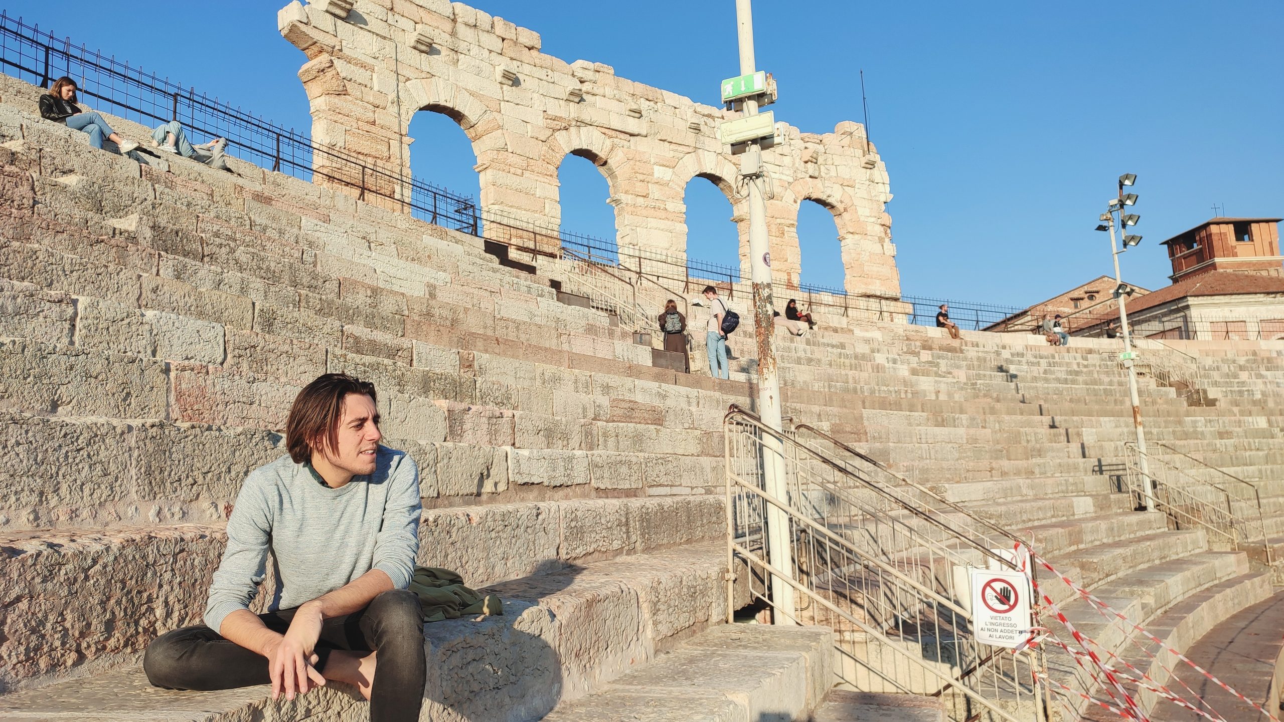 Persona sentada en los escalones de piedra de un antiguo anfiteatro a la luz del sol, con otras personas al fondo y tres grandes arcos de piedra visibles. | Jacob Warn