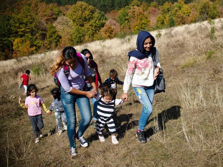 A diverse group of adults and children walk uphill through a grassy field surrounded by trees, hands held in unity as they journey across borders. | Jacob Warn