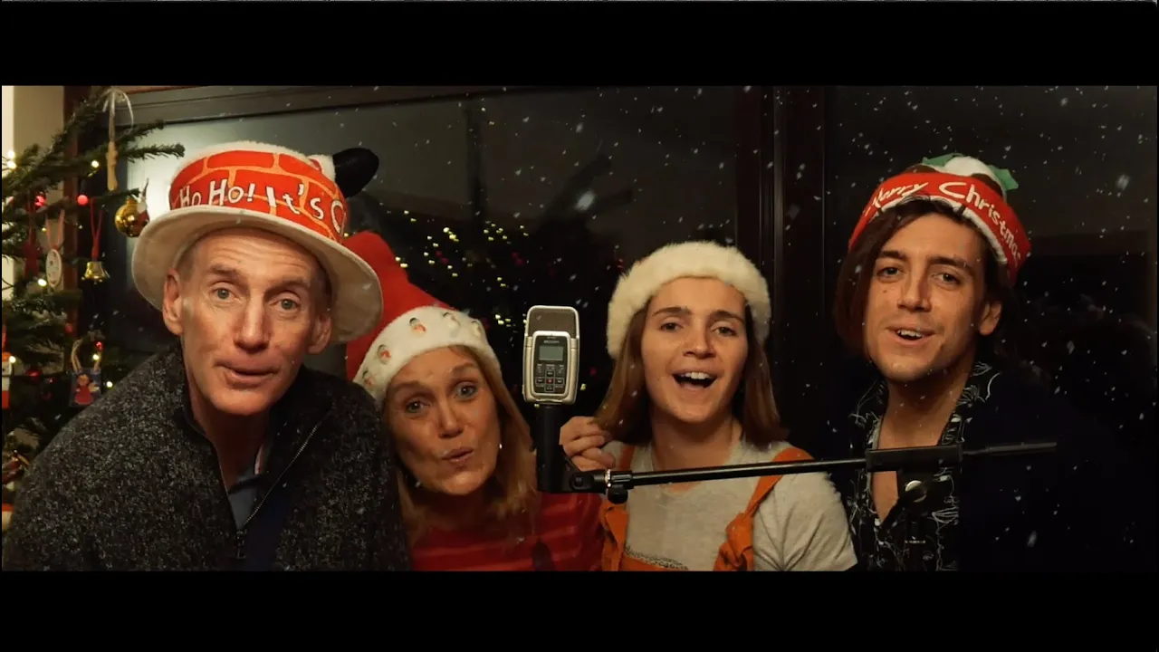 Cuatro miembros de una familia con gorros festivos cantan juntos "Only You" delante de un micrófono, con una ventana nevada y adornos navideños enmarcando su alegre actuación. | Jacob Warn