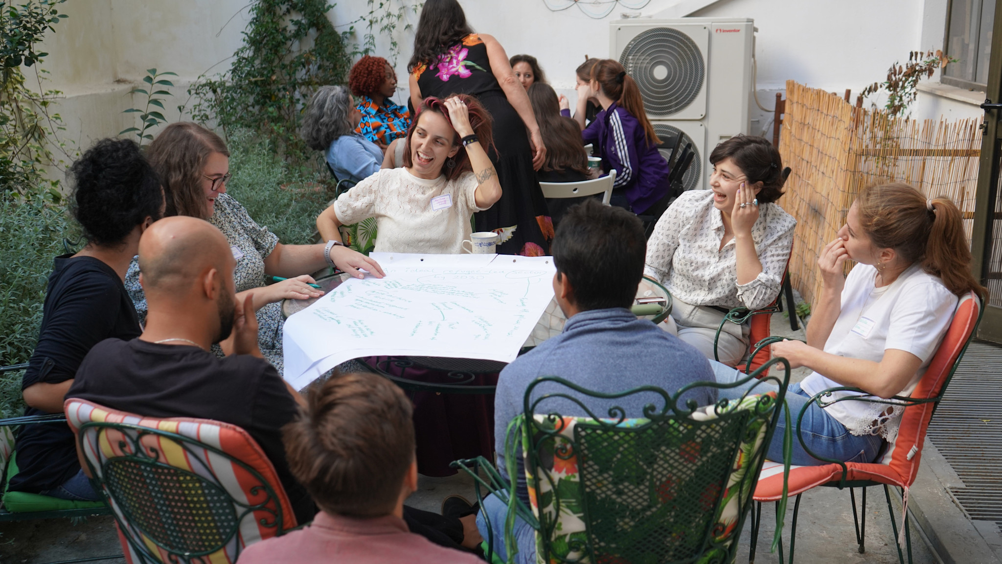 A group of people sits around a table outdoors, forming a community of practice, as they engage in discussion. One person points to a large sheet of paper in the center. | Jacob Warn